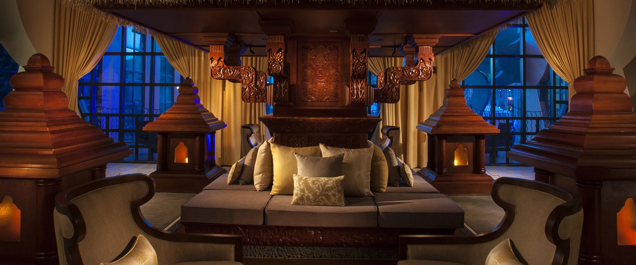 Luxurious, Balinese-inspired lounge with carved wooden details, a thatched canopy, plush seating, decorative pillows, and soft lighting, set against large windows with a twilight city view.