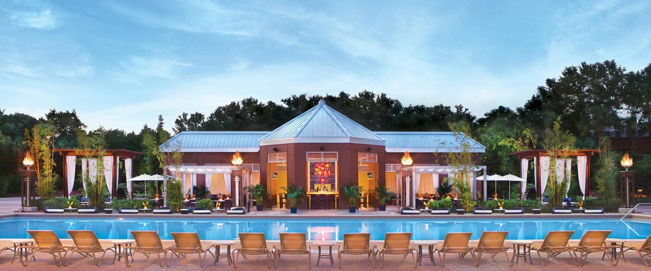 A row of lounge chairs faces a large outdoor swimming pool, with a modern pavilion and cabanas in the background, surrounded by greenery and lit torches at sunset.