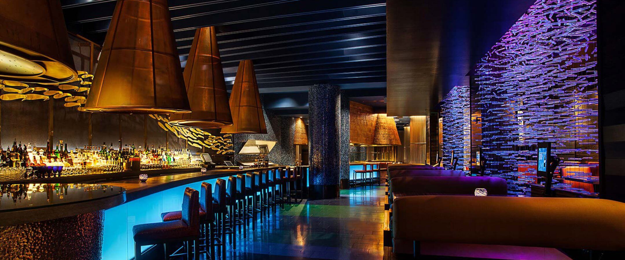 A modern, upscale bar with glowing blue lighting, copper cone-shaped light fixtures, high bar stools, and plush booth seating. The space features artistic wall designs and a sleek, colorful ambiance.