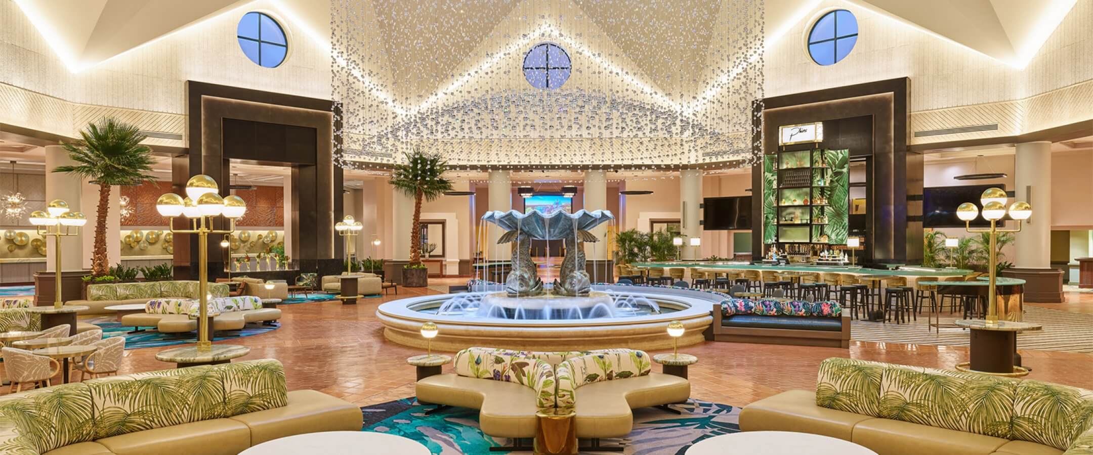Spacious, elegant hotel lobby with a central fountain, stylish seating areas, patterned rugs, lush plants, and high ceilings adorned with hanging lights and large round windows.
