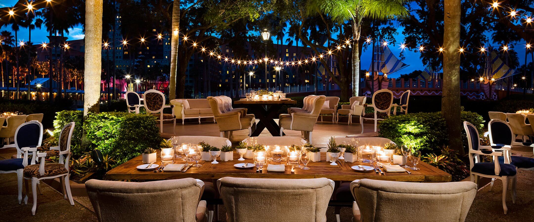 An elegant outdoor dining area at dusk features tables with candles, cushioned chairs, string lights overhead, and palm trees surrounding the space, creating a warm and inviting atmosphere.