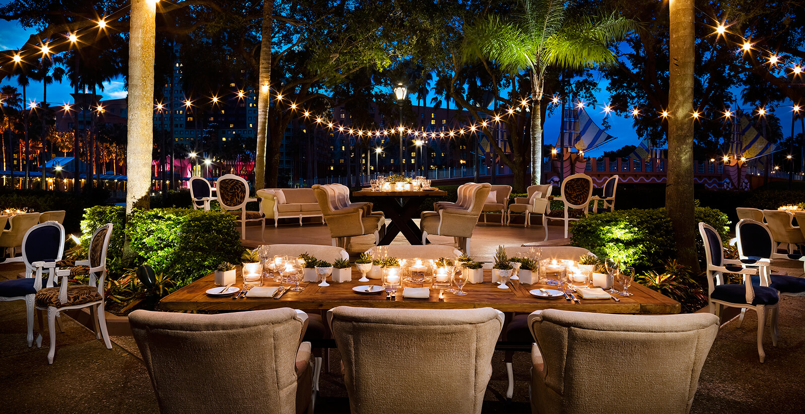 An elegant outdoor dining area at dusk features tables with candles, cushioned chairs, string lights overhead, and palm trees surrounding the space, creating a warm and inviting atmosphere.