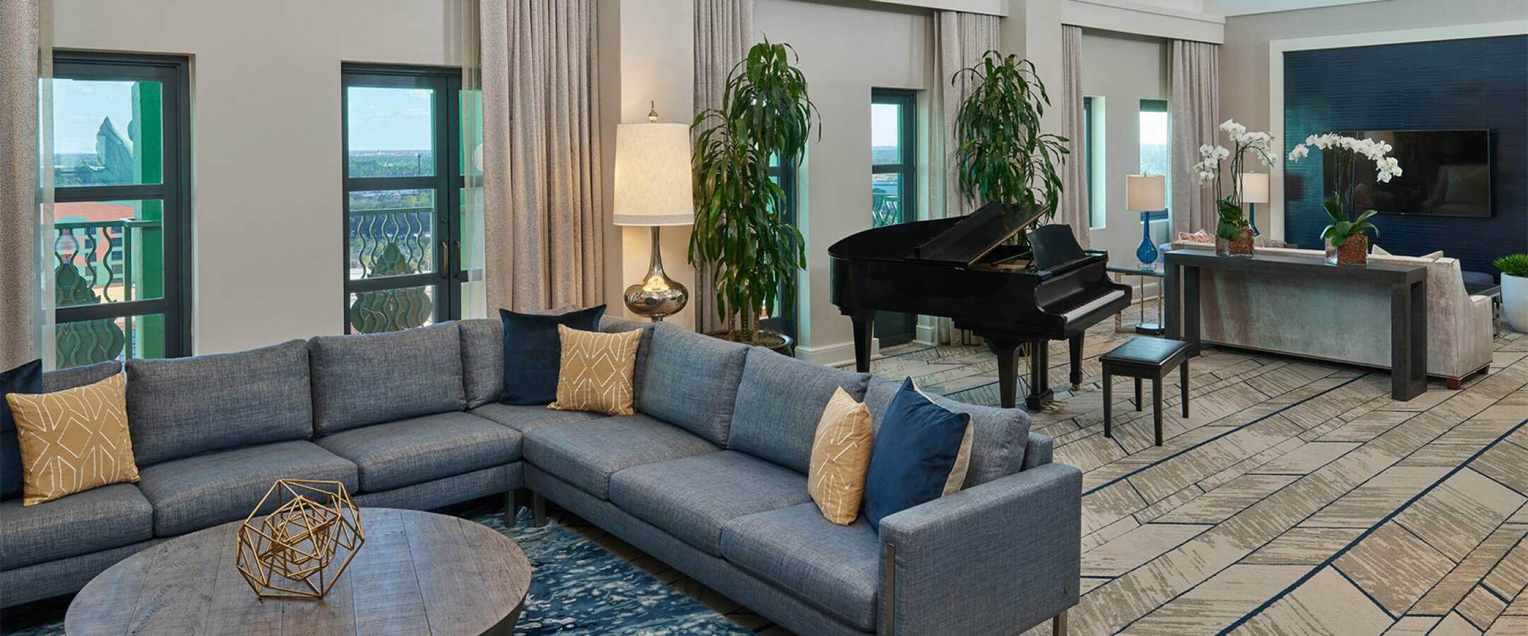 Modern living room with large sectional sofa, round wooden coffee table, grand piano, floor-to-ceiling windows, tall plants, and decorative accents. Light-filled space with contemporary decor and patterned carpet.