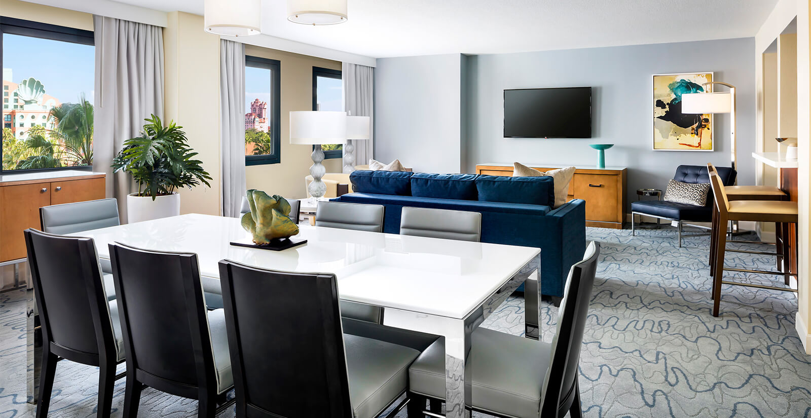 Modern hotel suite with a white dining table and black chairs, blue sofa, flat-screen TV, artwork on the wall, and large windows letting in natural light. Room is decorated with plants and contemporary furnishings.