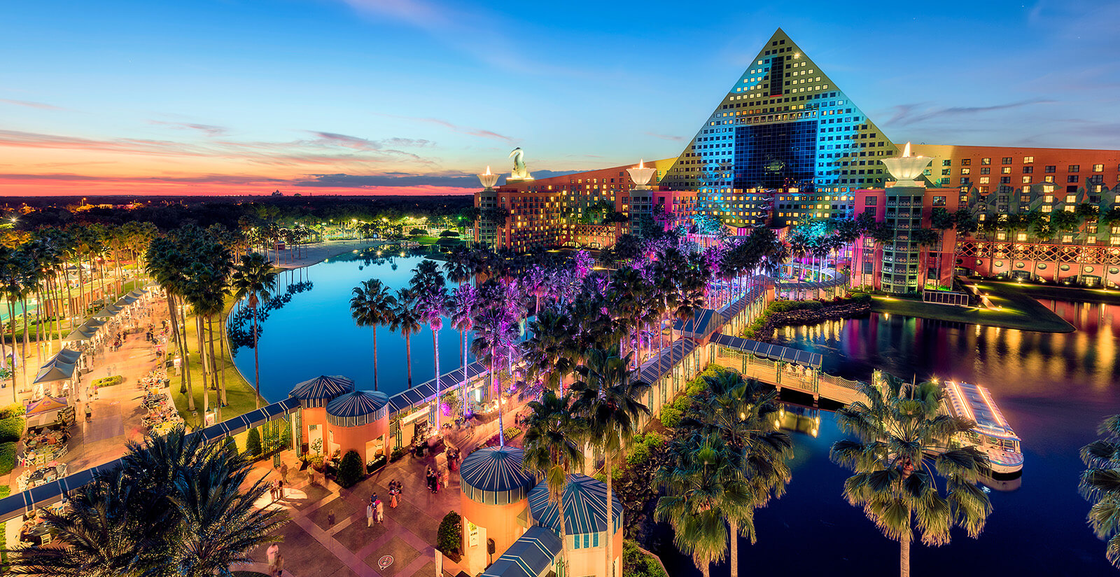 A vibrant, illuminated resort with a large triangular building, palm trees, a lake, pools, and pathways at sunset. The sky is colorful, reflecting on the water, creating a lively, tropical atmosphere.