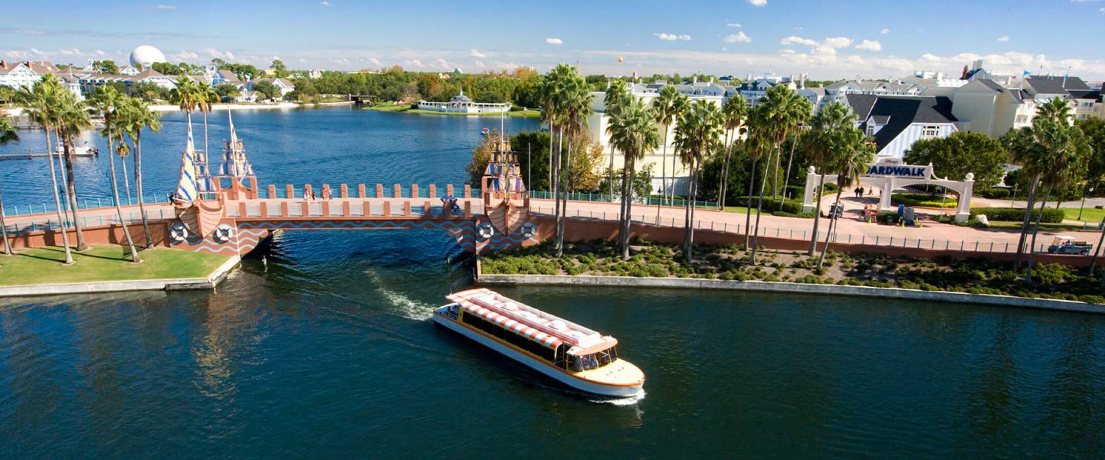 A boat travels along a canal near a decorative bridge, with palm trees and waterfront buildings in the background under a blue sky at a lively resort area.