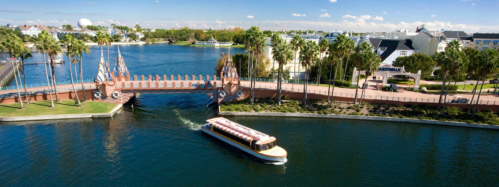 A boat travels along a canal near a decorative bridge, with palm trees and waterfront buildings in the background under a blue sky at a lively resort area.