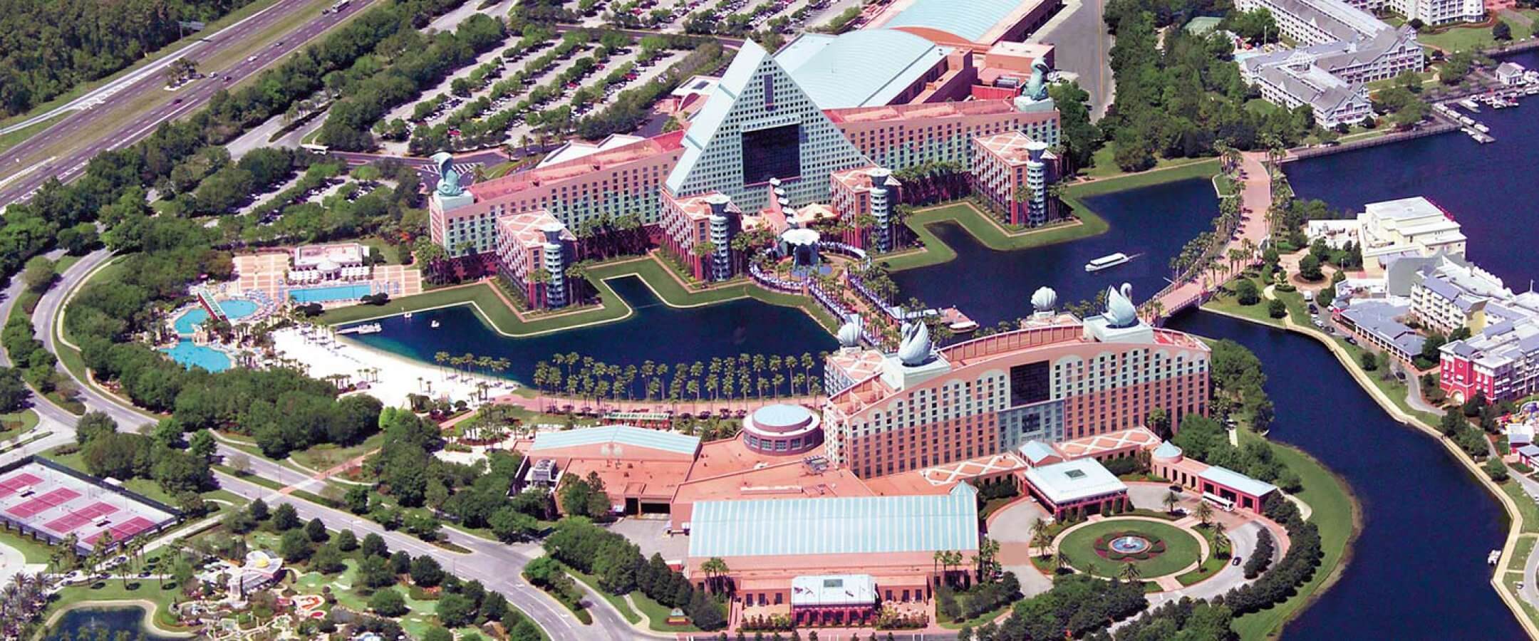 Aerial view of a large resort complex with colorful buildings, a pyramid-shaped structure, surrounding water features, pools, tennis courts, green spaces, and nearby roads and parking areas.