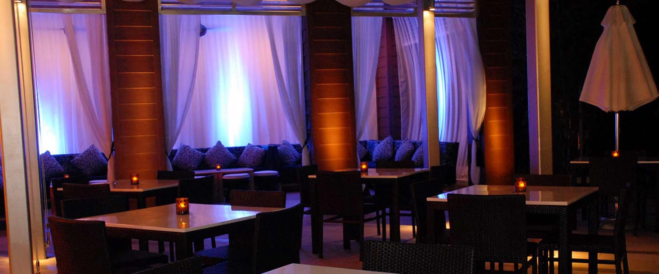 A modern outdoor lounge with dark wicker chairs, white tables, and glowing candle centerpieces. The background features draped white curtains lit by soft blue and purple lights, creating a relaxed, upscale ambiance.