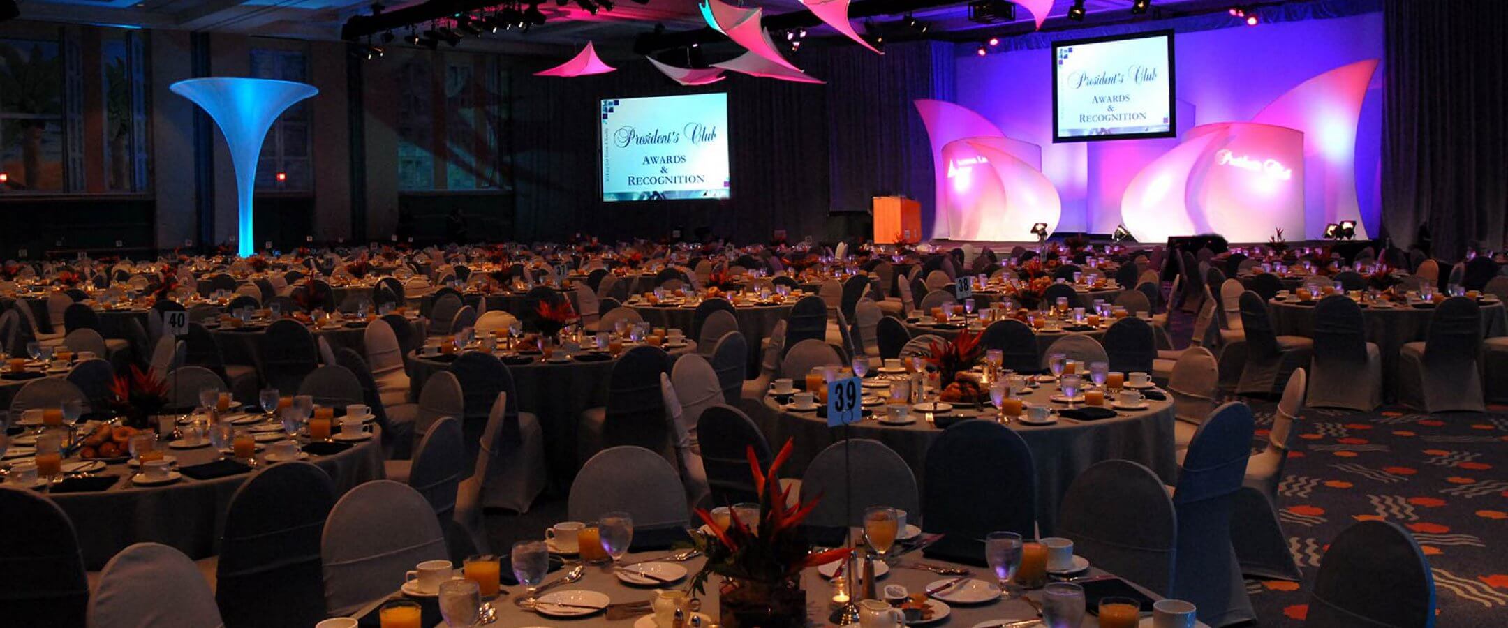A large banquet hall set up for an awards event, with round tables covered in white cloths, place settings, and breakfast food. The stage has pink lighting, large screens, and Awards Recognition displayed.