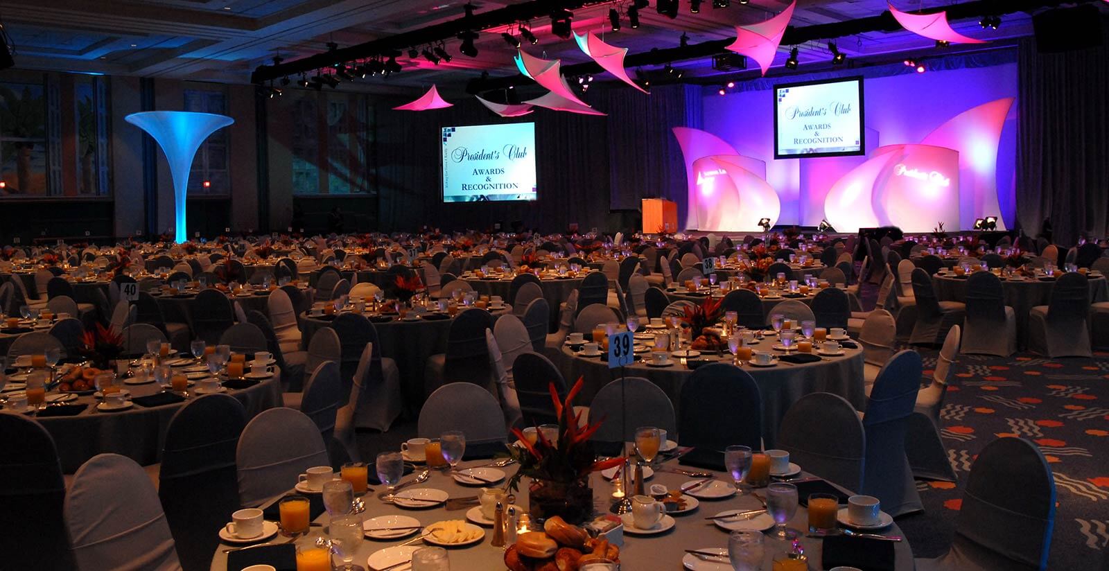 A large banquet hall set up for an awards event, with round tables covered in white cloths, place settings, and breakfast food. The stage has pink lighting, large screens, and Awards Recognition displayed.