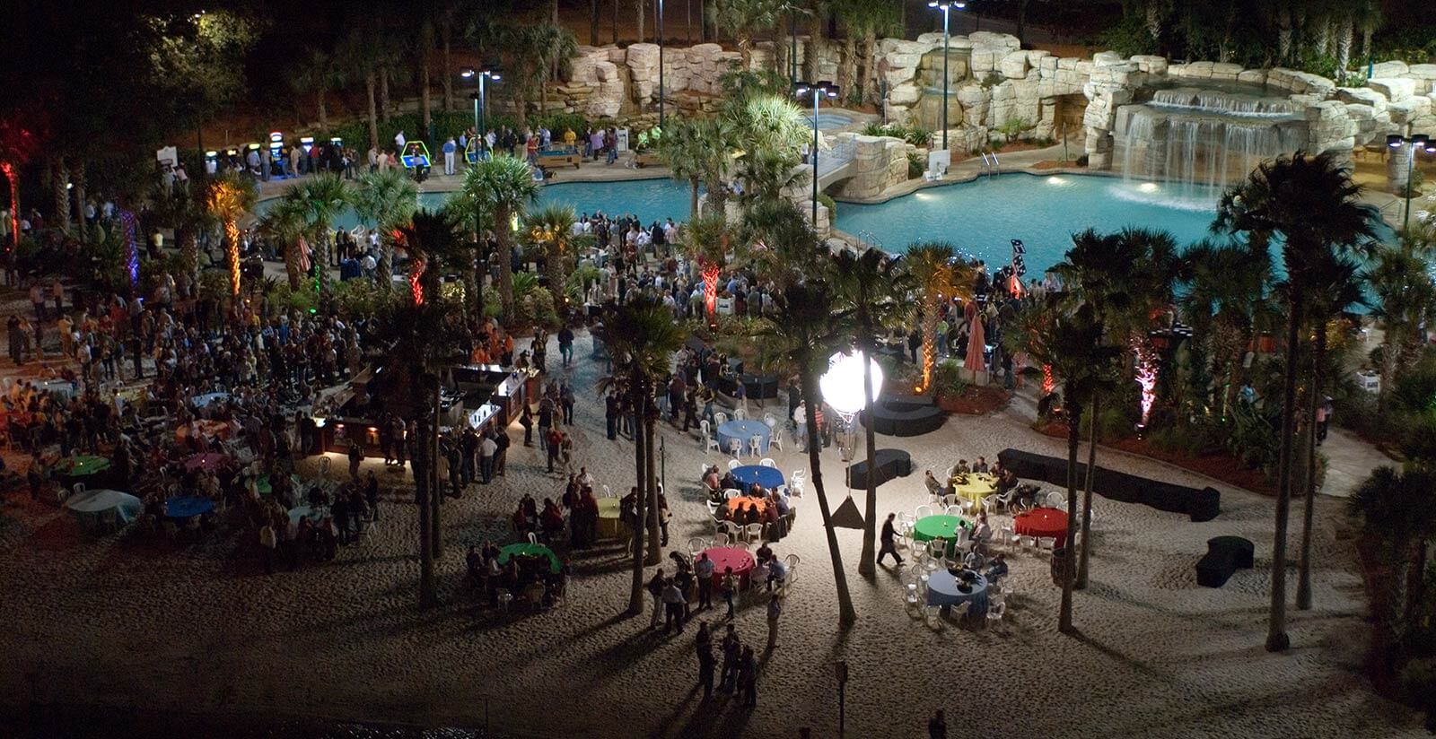A large nighttime outdoor event by a pool, with crowds of people, colorful round tables, palm trees, and bright lighting. A waterfall feature is visible in the background, creating a lively, festive atmosphere.
