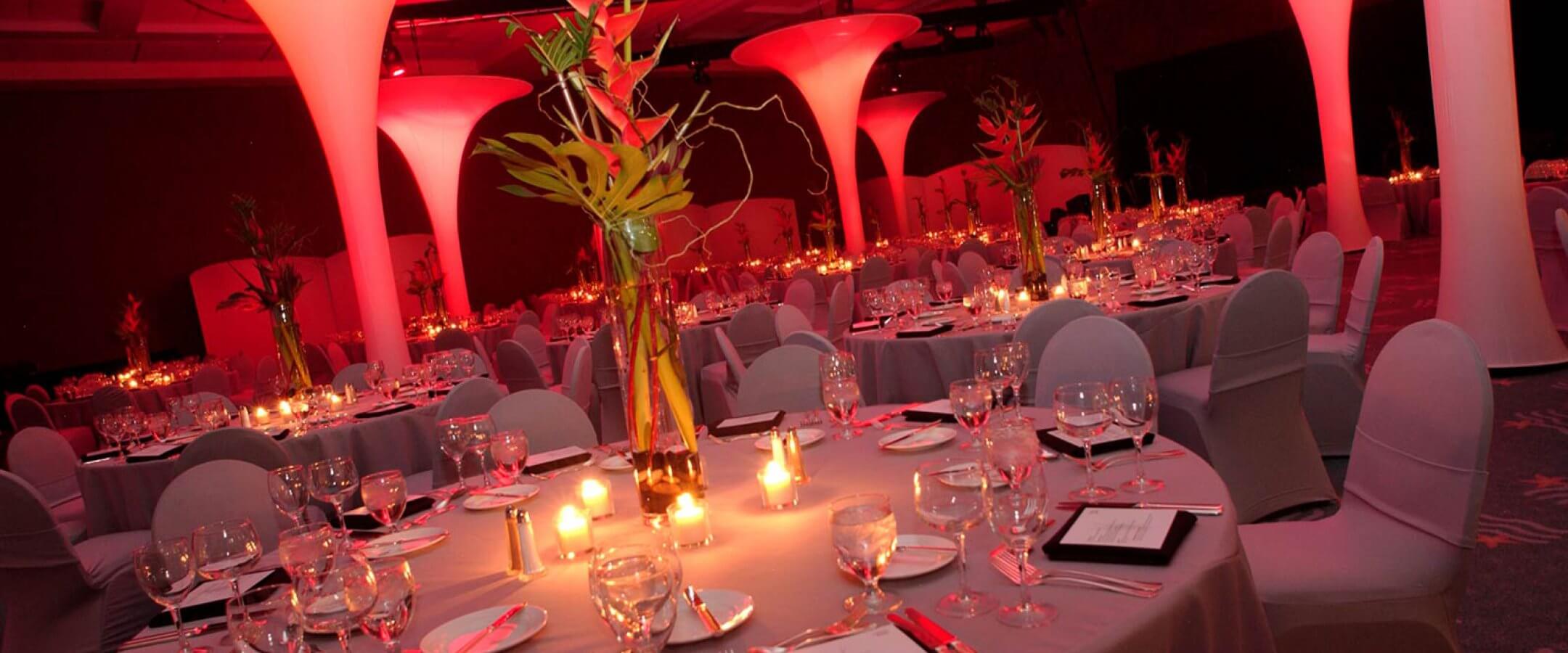 Elegant banquet hall with round tables set for dinner, decorated with candles and tall glass vases with green stems. Ambient pink lighting and large lighted pillars create a warm, festive atmosphere.