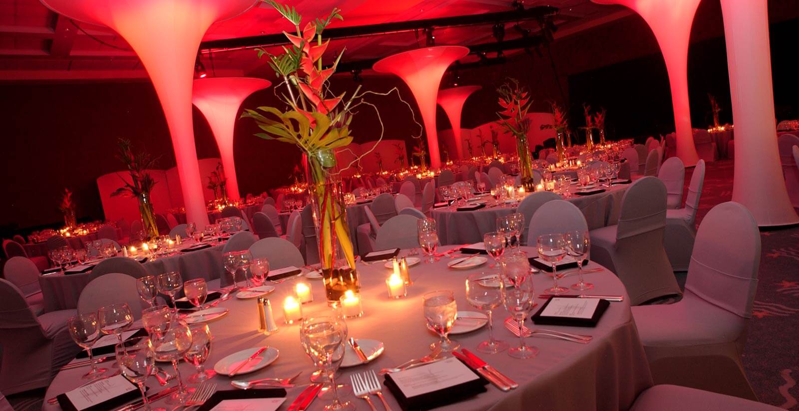 Elegant banquet hall with round tables set for dinner, decorated with candles and tall glass vases with green stems. Ambient pink lighting and large lighted pillars create a warm, festive atmosphere.