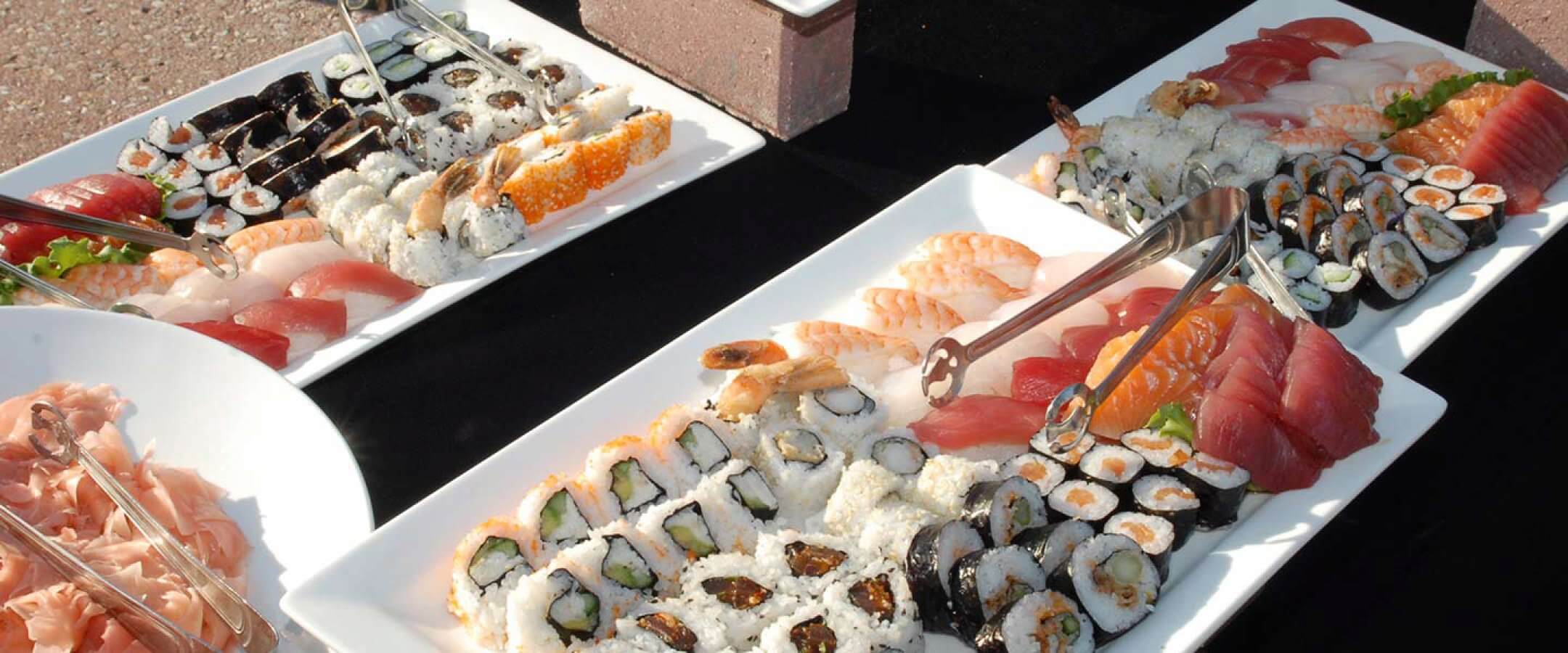 Plates filled with assorted sushi, including nigiri, sashimi, and various sushi rolls. Metal tongs rest on the plates, which are arranged on a black tablecloth. Ginger slices are visible on one plate.