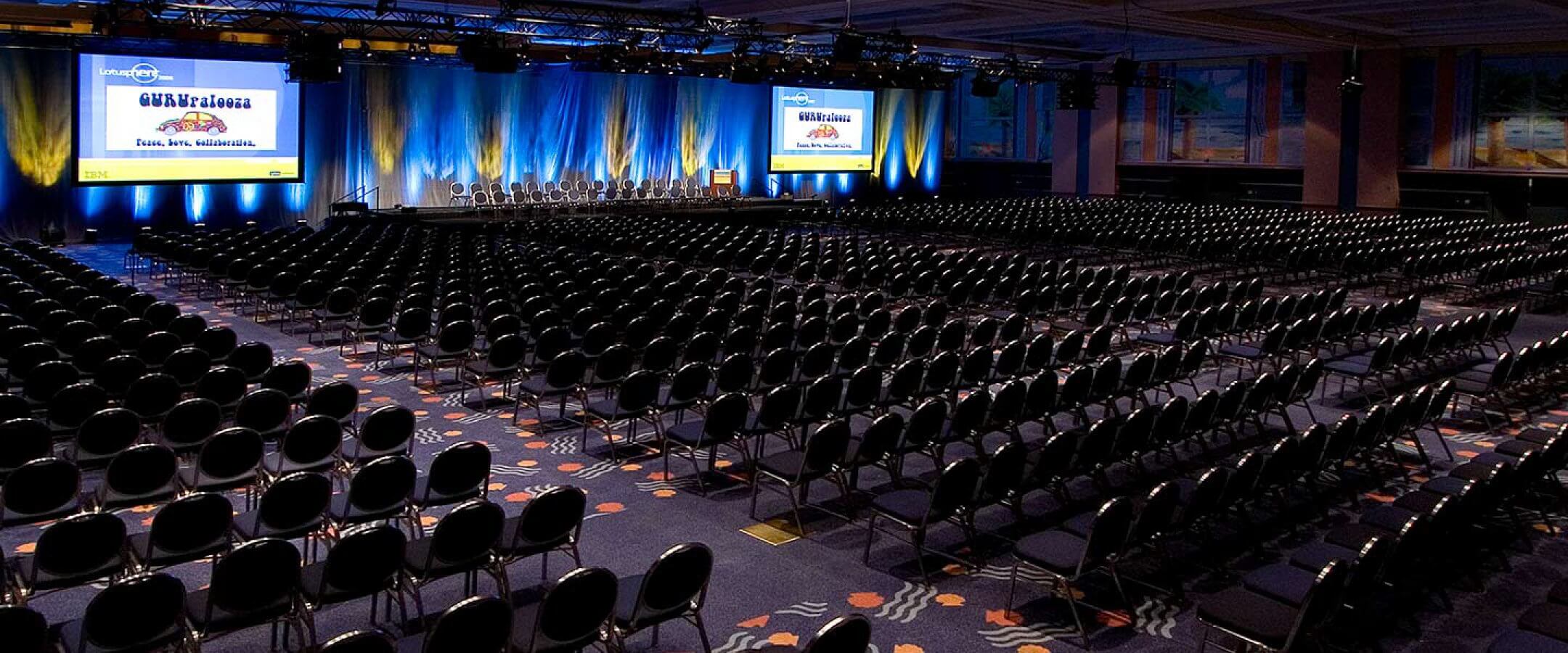 A large, empty conference hall with rows of black chairs facing a stage with a podium and two large projection screens displaying conference information, illuminated by blue and yellow lights.