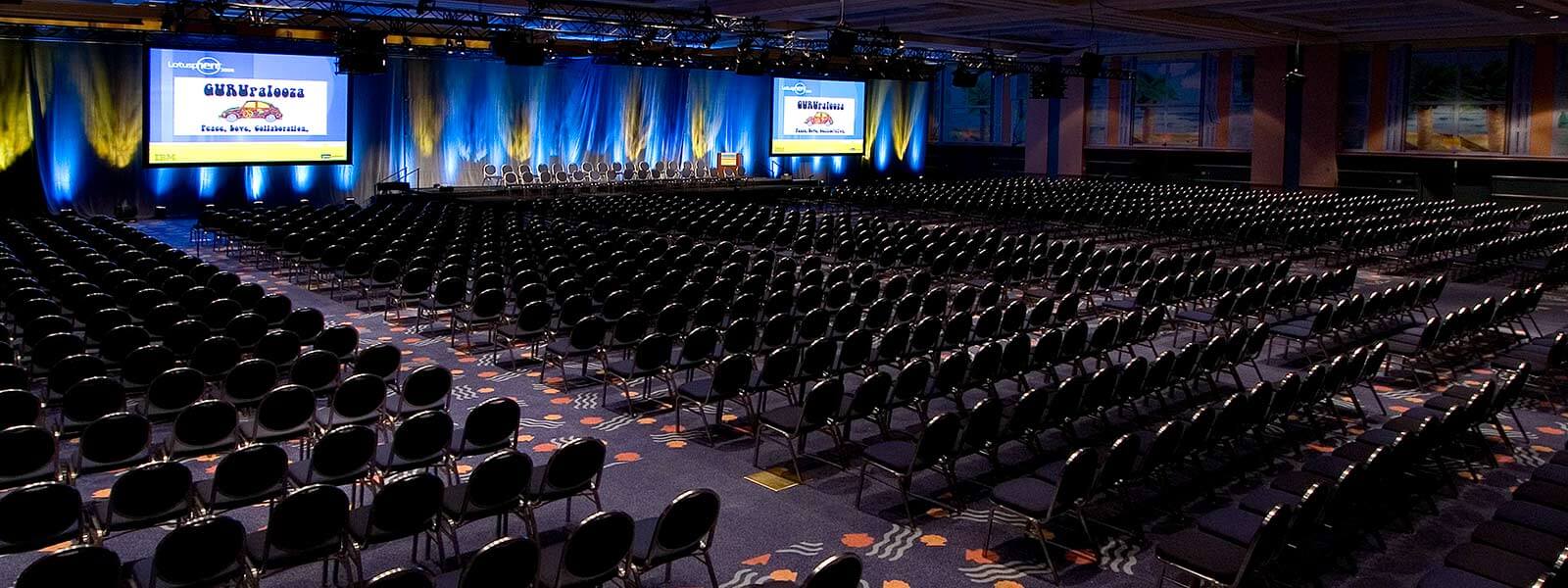 A large, empty conference hall with rows of black chairs facing a stage with a podium and two large projection screens displaying conference information, illuminated by blue and yellow lights.