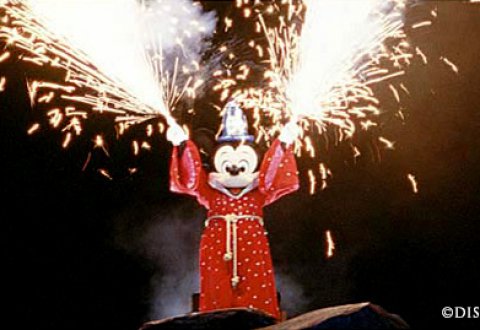 Mickey Mouse dressed as a sorcerer stands on a rock at night, raising both arms as fireworks and sparks shoot from his hands.