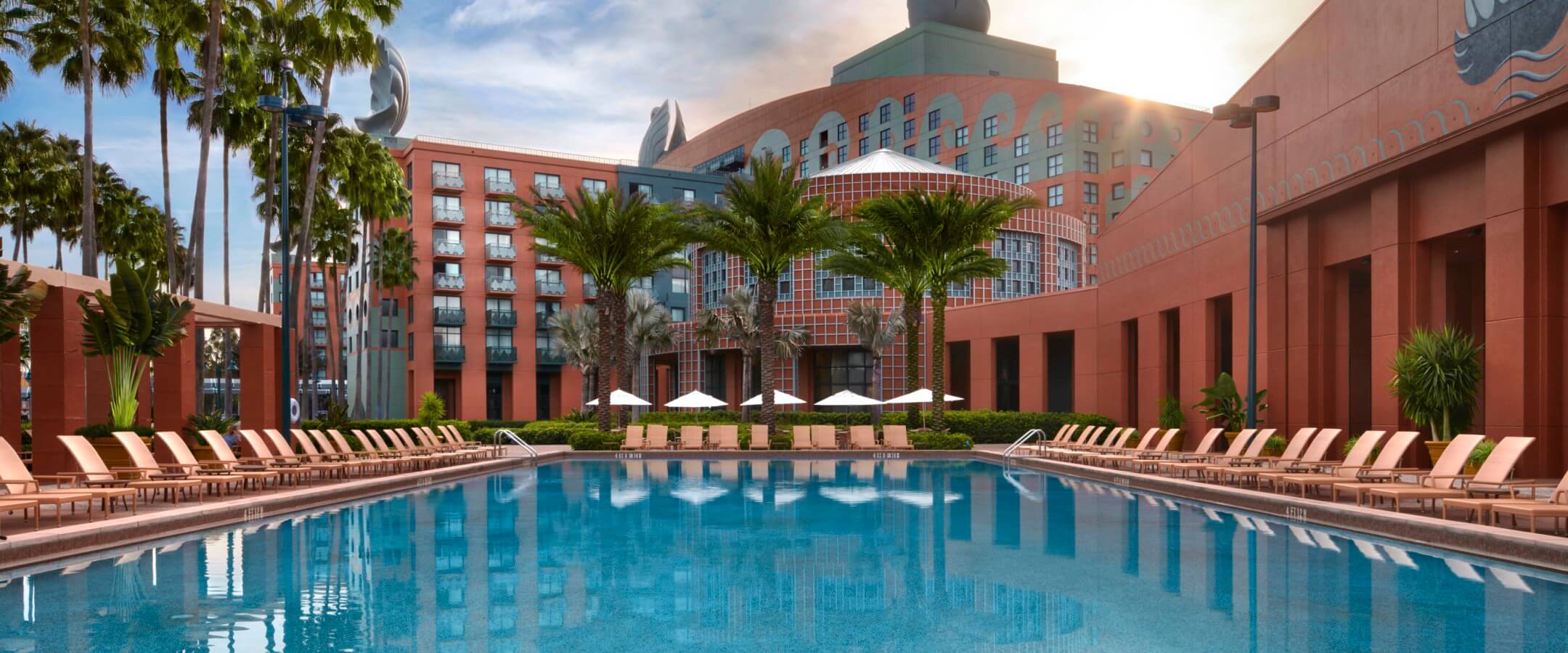 Large outdoor swimming pool surrounded by lounge chairs and palm trees, with a distinctive hotel building featuring swan statues and a blue sky in the background.
