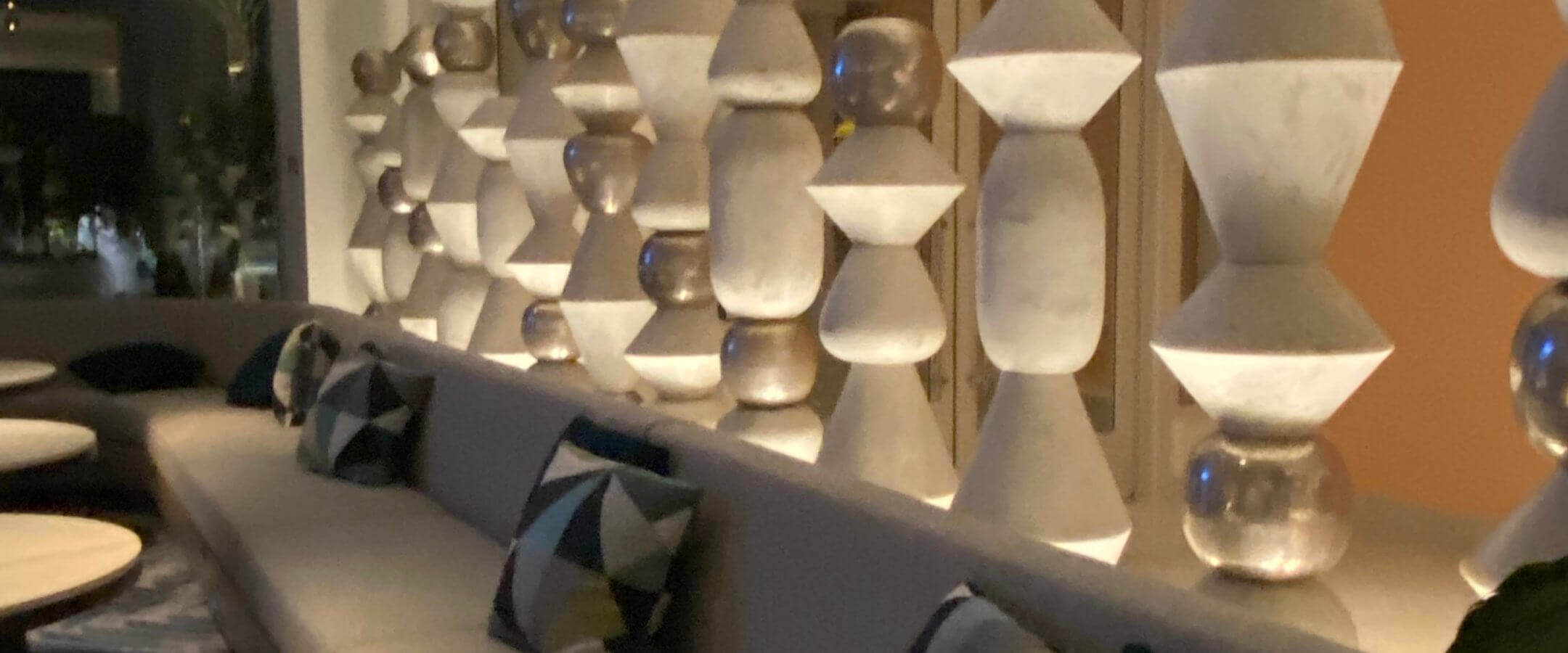 A modern lounge area with a long cushioned bench, geometric-patterned pillows, round white tables, and a decorative partition featuring stacked hourglass-shaped sculptures, softly lit by ambient lighting.