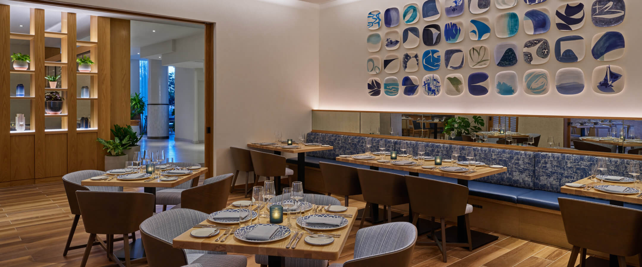 Modern restaurant dining area with wooden floors, brown chairs, set tables, a blue-patterned cushioned bench, and a wall decorated with square ceramic art pieces in blue and white tones. Warm, ambient lighting throughout.
