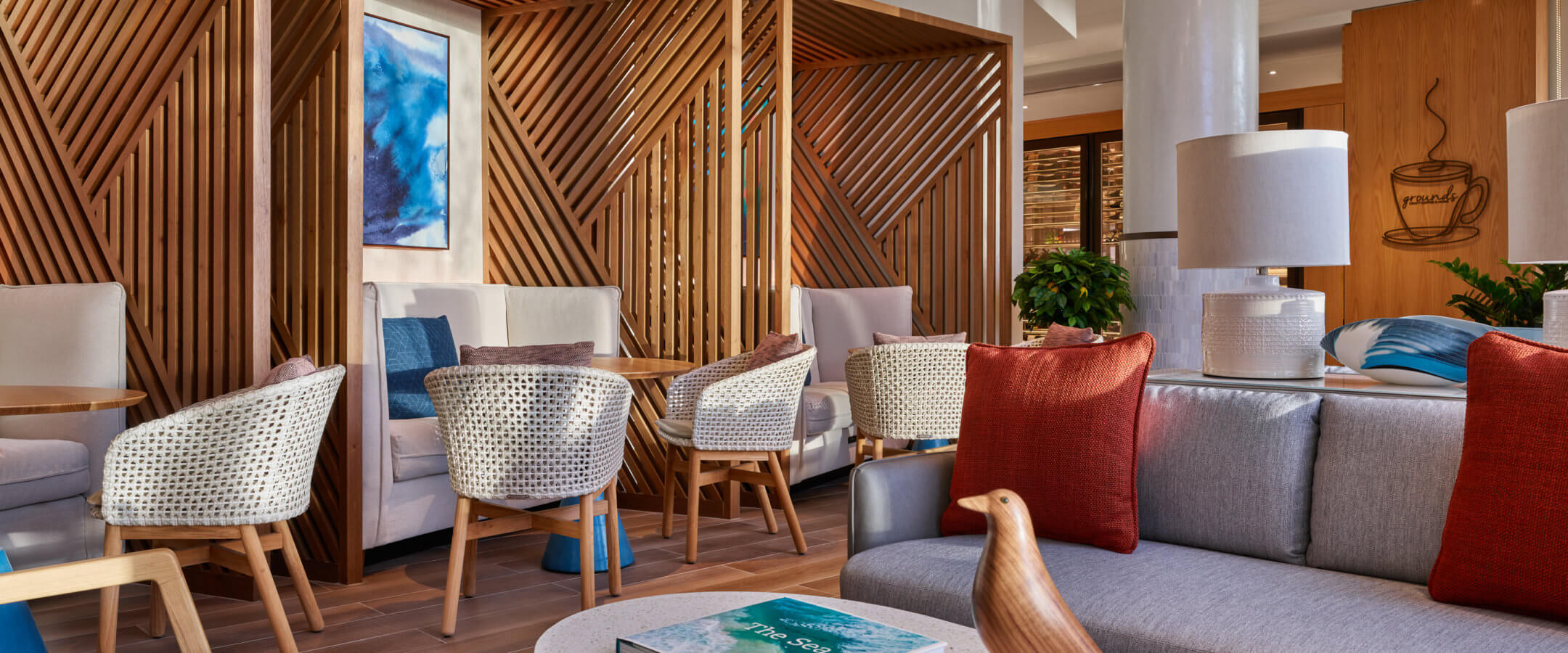 Modern lounge area with wood accents, cozy armchairs, a gray sofa with red pillows, a coffee table with a decorative bird and book, and wall art with blue tones. The space is inviting and well-lit.