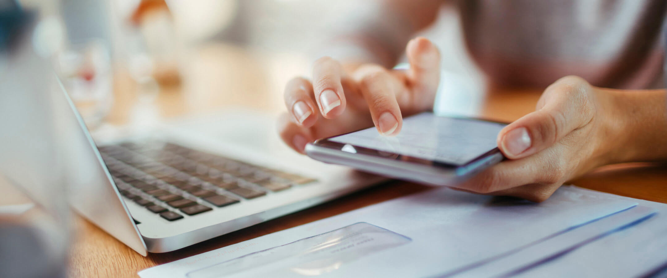 A person uses a smartphone with one hand while the other hand rests on a laptop keyboard. Papers and an envelope are on the table, suggesting multitasking or online work.
