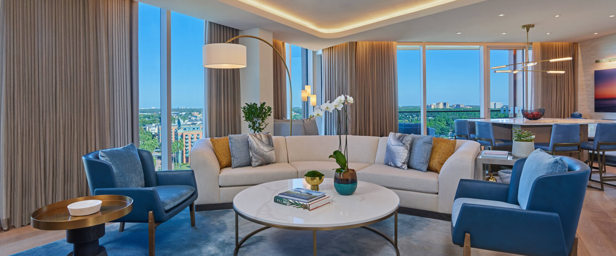 A modern, bright living room with floor-to-ceiling windows, a curved white sofa, blue armchairs, a round coffee table, and a blue rug. City views are visible outside, and a dining area is in the background.