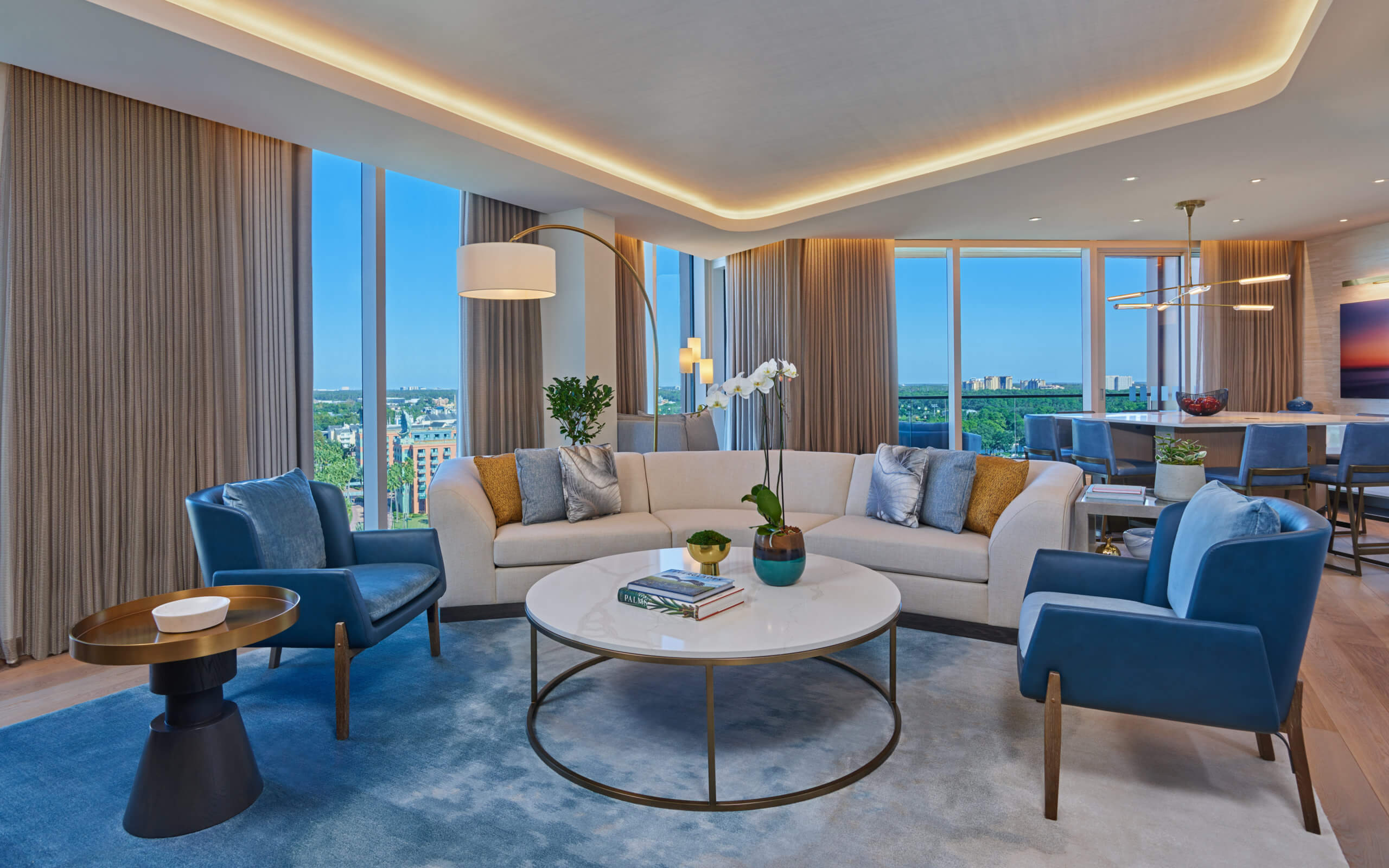 A modern, bright living room with floor-to-ceiling windows, a curved white sofa, blue armchairs, a round coffee table, and a blue rug. City views are visible outside, and a dining area is in the background.
