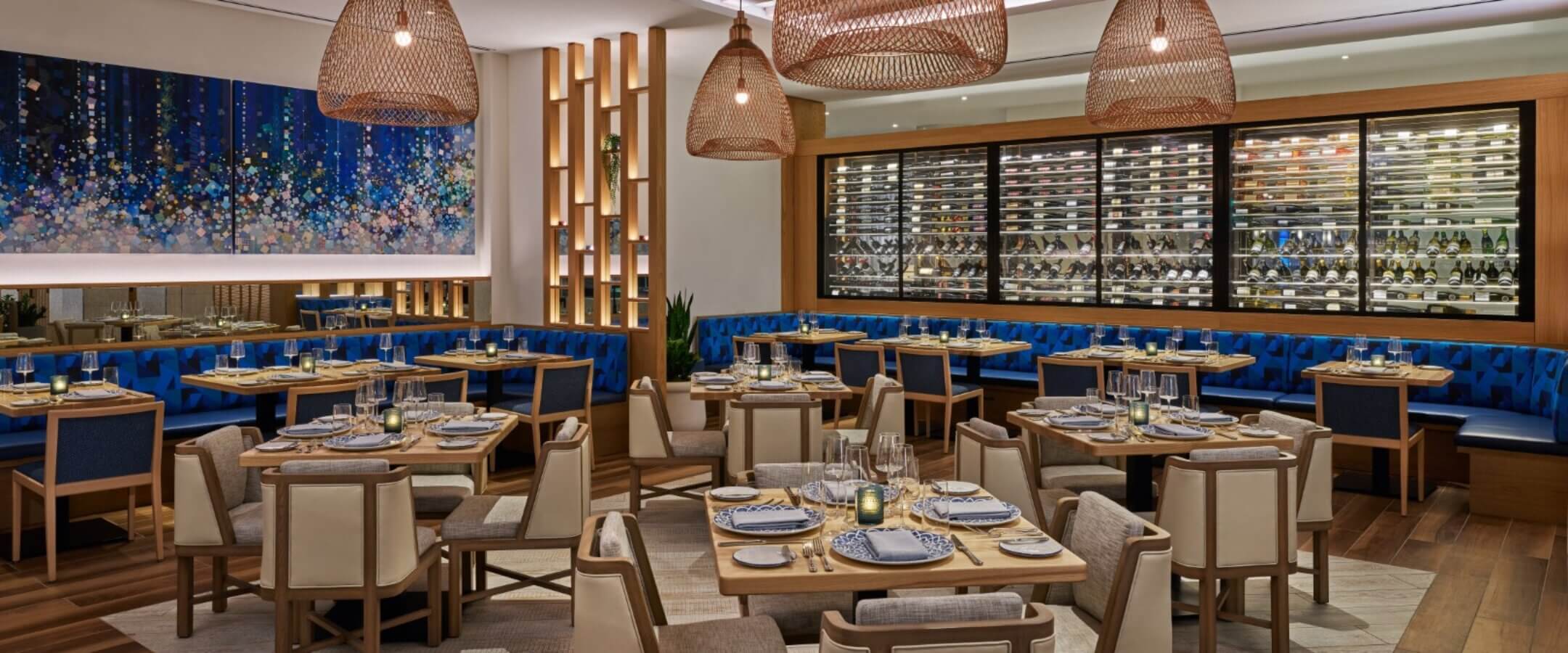 Elegant restaurant interior with wooden tables and cushioned chairs, blue upholstered seating along the walls, modern pendant lights, a wall of wine bottles, and a large abstract painting.