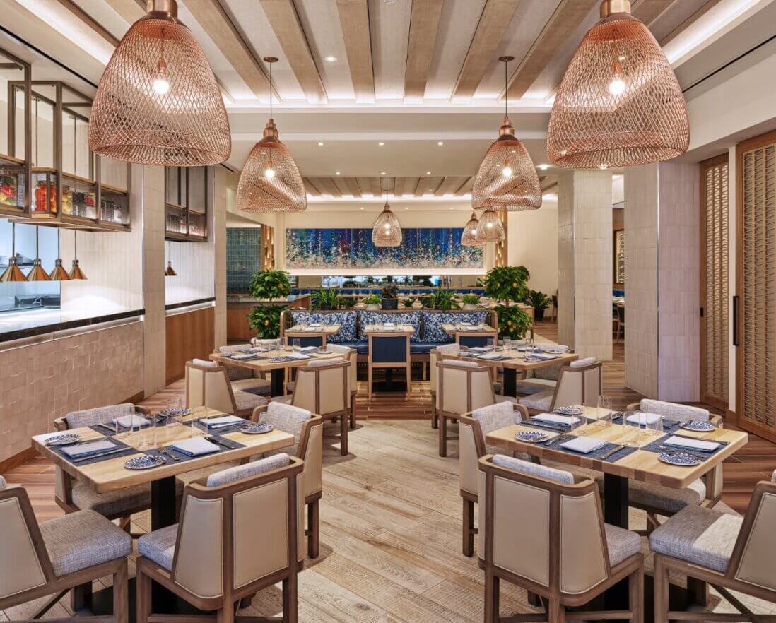 A modern, elegant restaurant interior with wooden tables, cushioned chairs, woven pendant lights, and decorative blue tiles on the back wall. Plants and soft lighting create a warm, inviting atmosphere.
