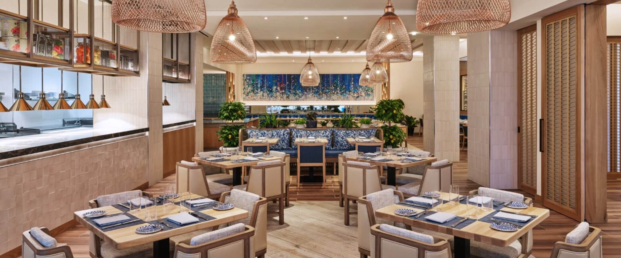 A modern, elegant restaurant interior with wooden tables, cushioned chairs, woven pendant lights, and decorative blue tiles on the back wall. Plants and soft lighting create a warm, inviting atmosphere.
