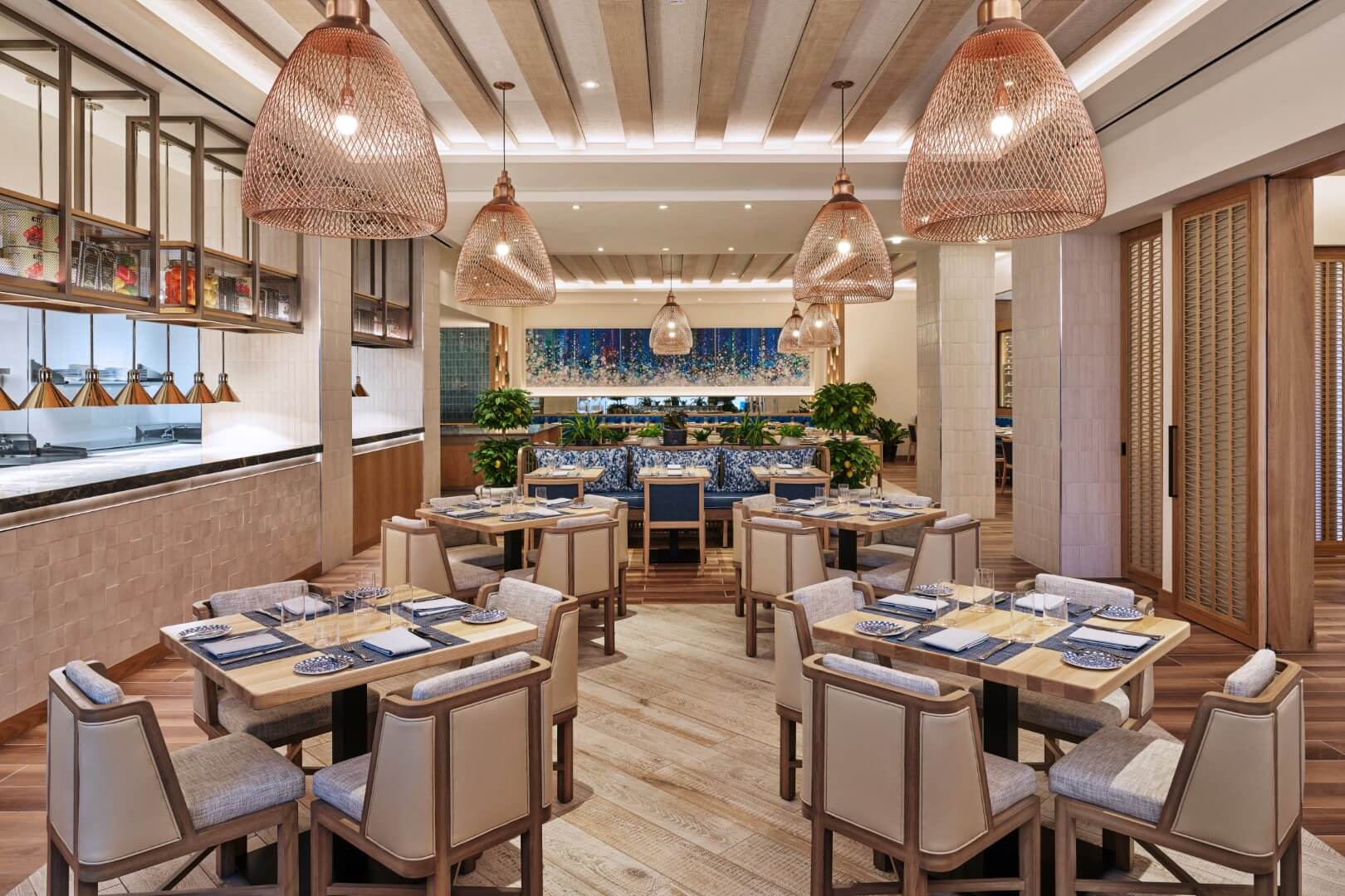 Um interior de restaurante moderno e elegante com mesas de madeira, cadeiras almofadadas, luminárias pendentes de tecido e azulejos azuis decorativos na parede do fundo. As plantas e a iluminação suave criam uma atmosfera calorosa e convidativa.