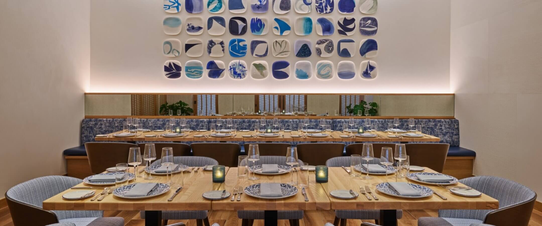 A modern dining room with a long wooden table set with plates, glasses, and napkins. Plush chairs surround the table. The back wall features a display of decorative blue and white plates arranged in a grid.