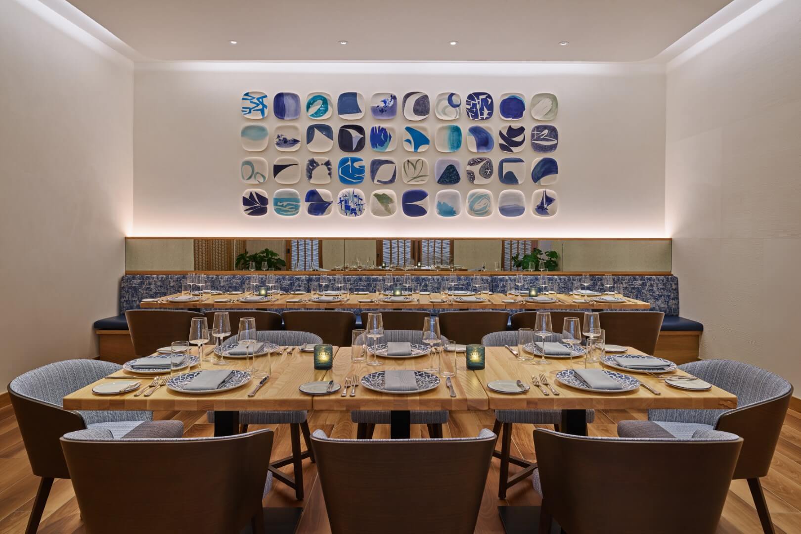 Uma sala de jantar moderna com uma longa mesa de madeira com pratos, copos e guardanapos. Cadeiras de pelúcia cercam a mesa. A parede do fundo apresenta uma exibição de pratos decorativos azuis e brancos dispostos em uma grade.