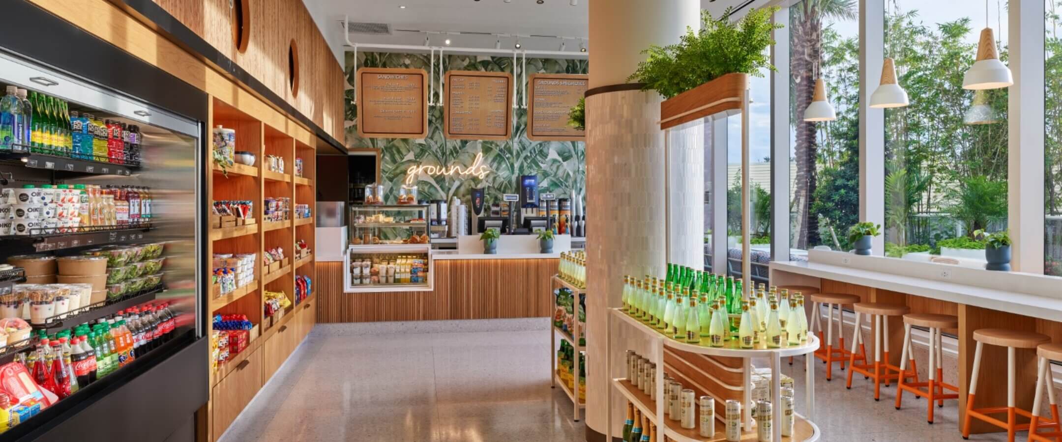 Bright, modern café with large windows, wooden shelves stocked with snacks and drinks, a curved counter with beverages, high stools by the window, and a leafy plant wall behind the coffee counter.