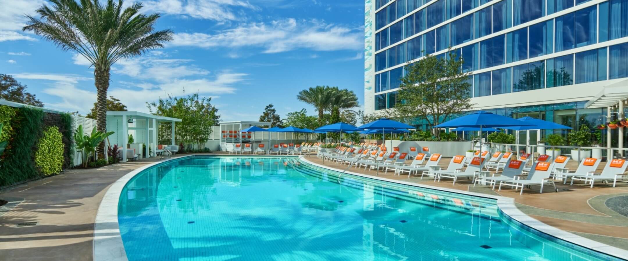 A curved outdoor swimming pool surrounded by orange and white lounge chairs, blue umbrellas, palm trees, and a modern glass hotel building under a sunny blue sky.