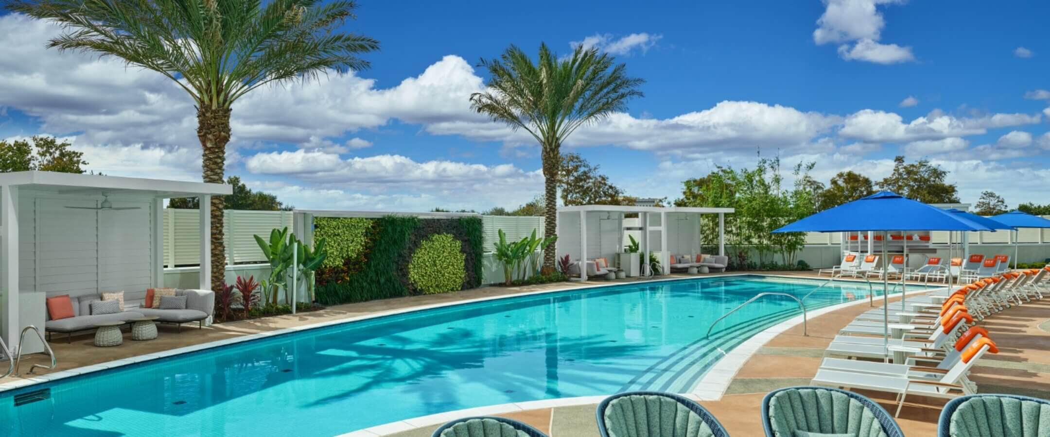 A modern outdoor pool area with clear blue water, surrounded by lounge chairs, orange and white accents, palm trees, shaded cabanas, and green plants under a bright, partly cloudy sky.