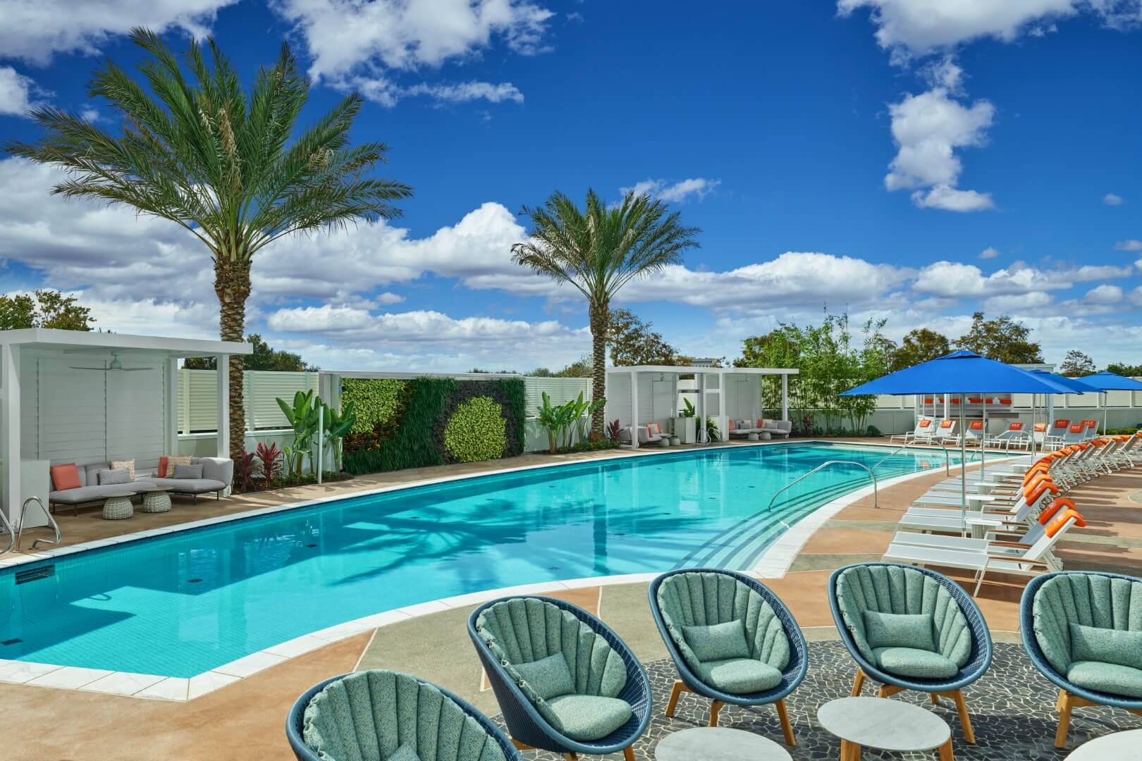 Una moderna area piscina all'aperto con acqua cristallina, circondata da sedie lounge , accenti arancioni e bianchi, palme, cabine ombreggiate e piante verdi sotto un cielo luminoso e parzialmente nuvoloso.