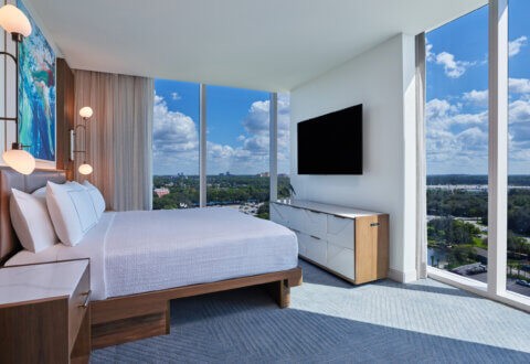 Modern hotel room with a large bed, bedside tables, flat-screen TV, dresser, and floor-to-ceiling windows offering a scenic view of a city and blue sky with clouds.