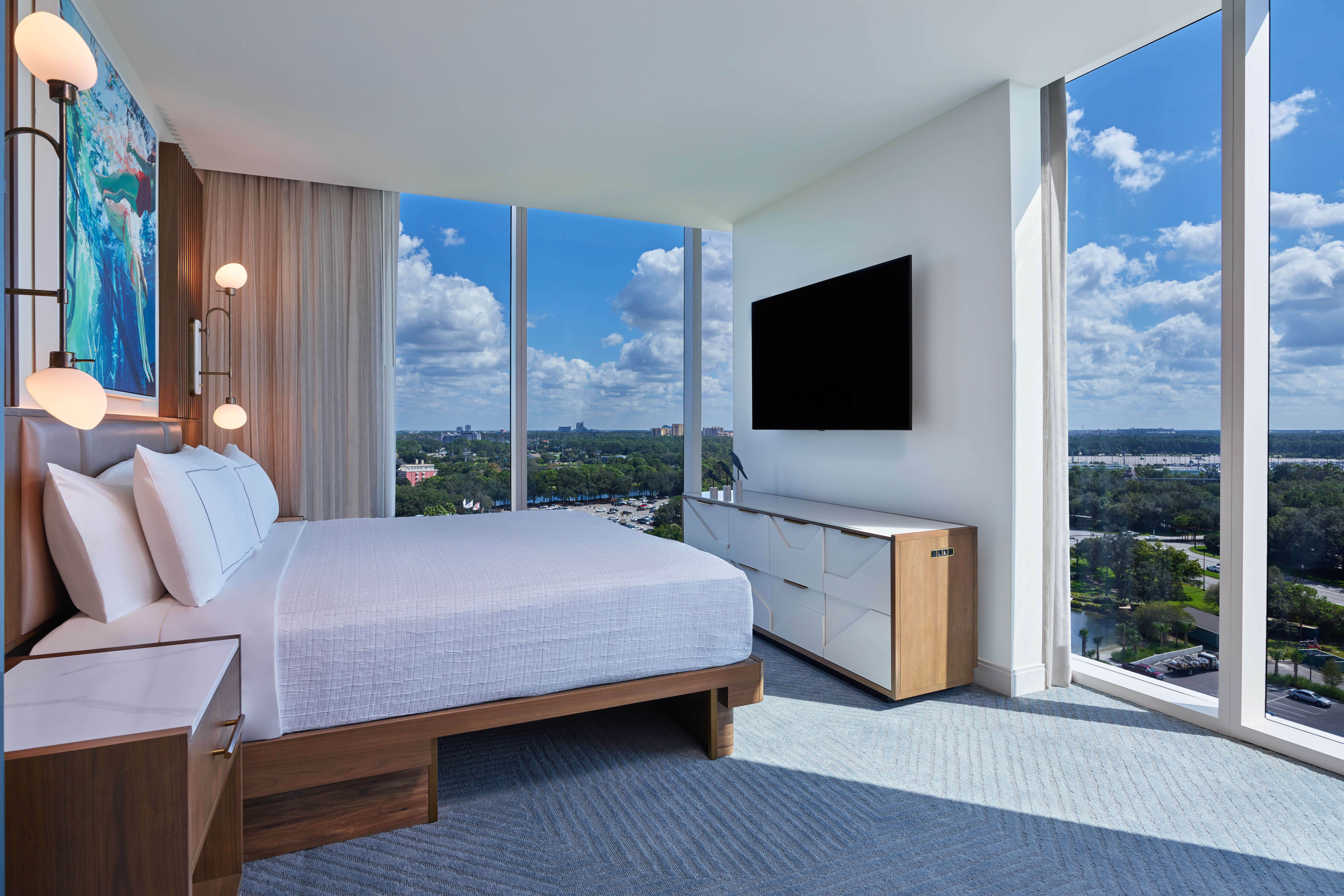 Modern hotel room with a large bed, abstract art, wall-mounted TV, floor-to-ceiling windows, and a scenic view of trees and sky. Sunlight fills the bright, airy space with minimalist decor.