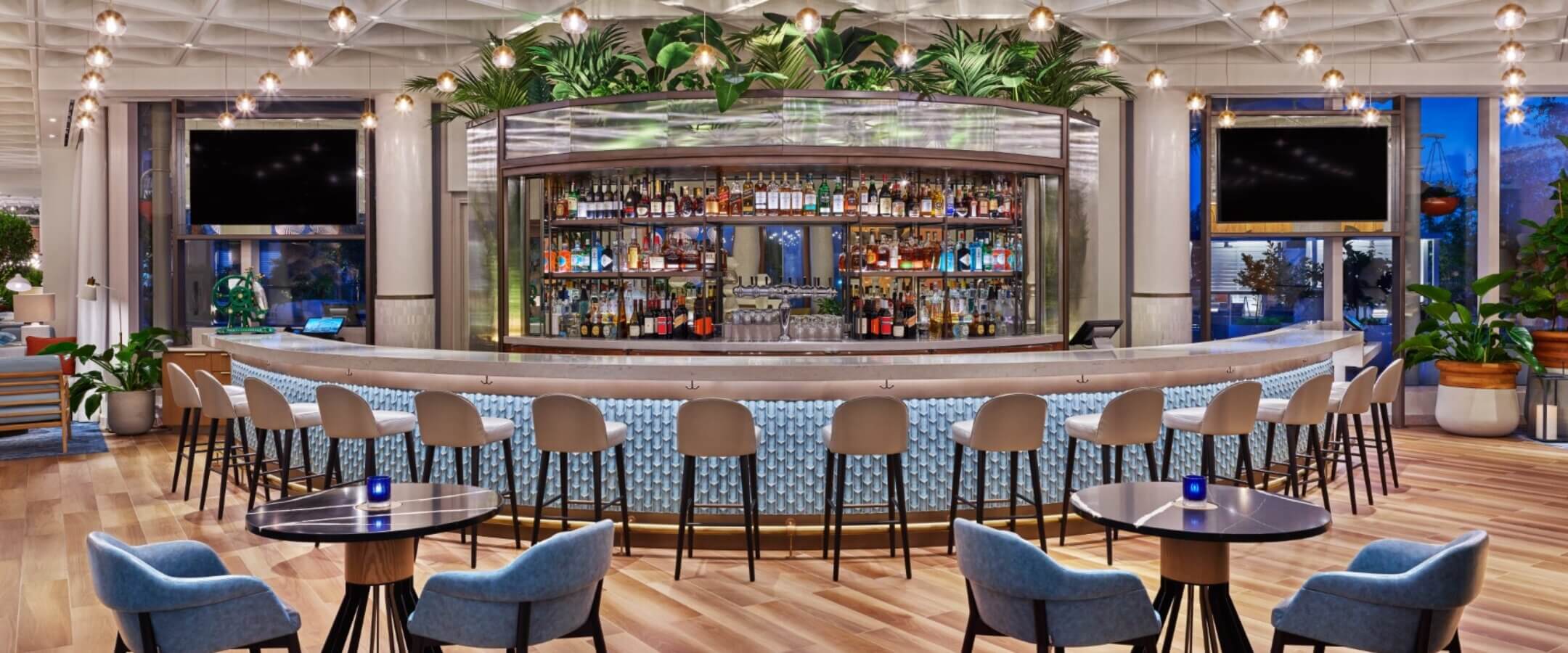 A modern, stylish bar with a curved counter, high stools, and small round tables with blue chairs. The bar is well-stocked, surrounded by greenery, and features a geometric ceiling with pendant lights.