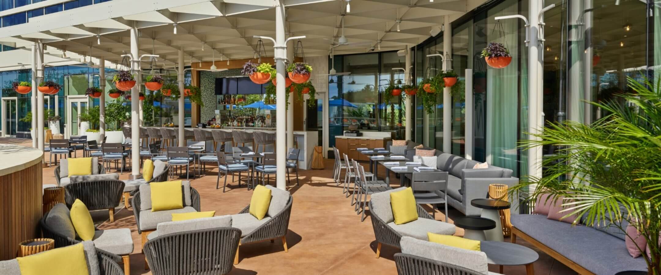 Outdoor restaurant patio with modern gray wicker chairs, yellow cushions, small tables, hanging potted plants, and a shaded bar area, situated next to a glass building on a sunny day.
