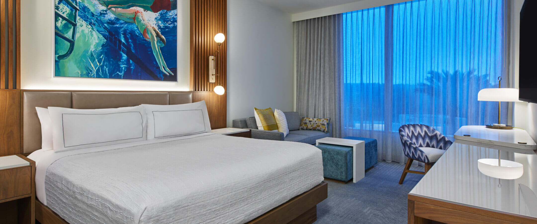 Modern hotel room with a large bed, contemporary furniture, colorful underwater-themed artwork above the bed, a sitting area by the window, and blue light filtering through sheer curtains.