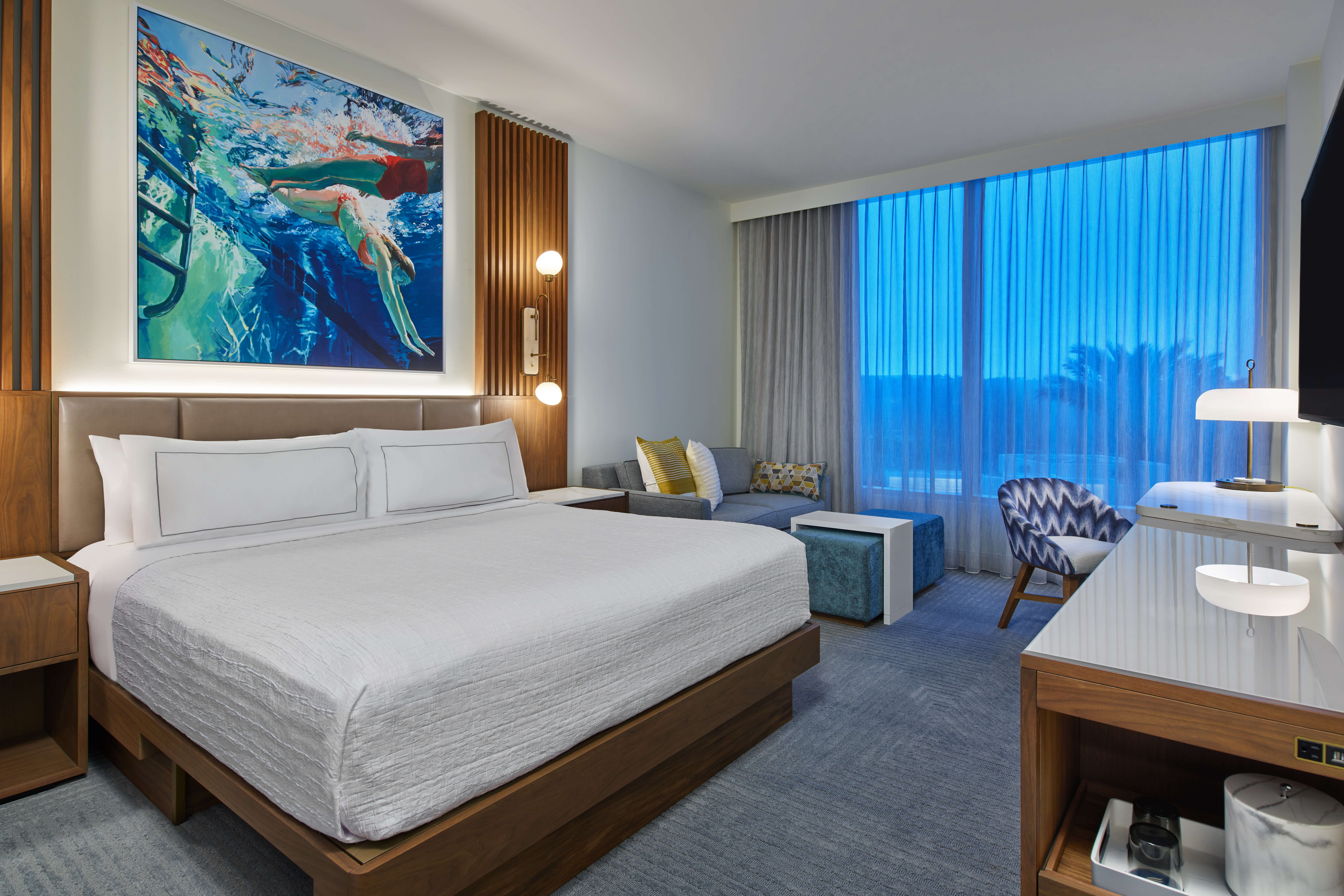 Modern hotel room with a large bed, contemporary furniture, colorful underwater-themed artwork above the bed, a sitting area by the window, and blue light filtering through sheer curtains.