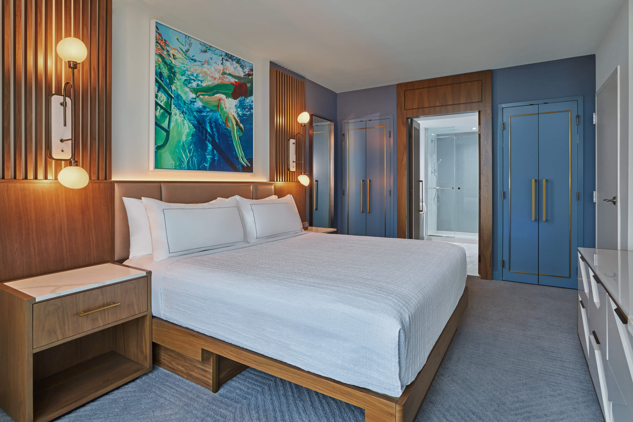 Modern hotel room with a large bed, white bedding, wooden headboard, two bedside tables, abstract surfer artwork above the bed, blue accent walls, and a view into a bathroom with a glass shower.