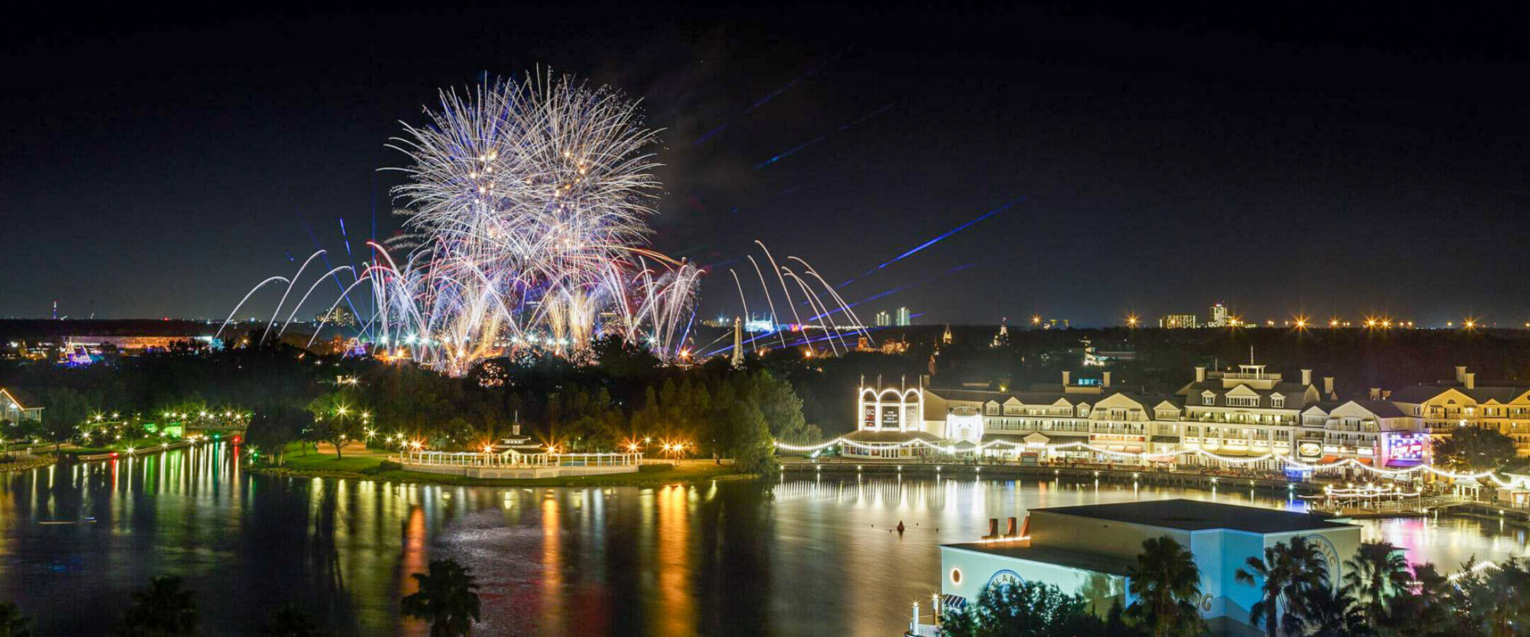 Fireworks explode in the night sky above a waterfront resort area, with brightly lit buildings reflecting off the calm water and a city skyline visible in the distance.