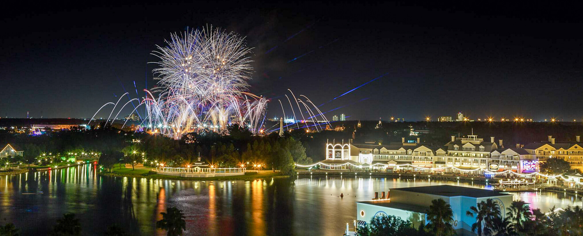 Fireworks explode in the night sky above a waterfront resort area, with brightly lit buildings reflecting off the calm water and a city skyline visible in the distance.