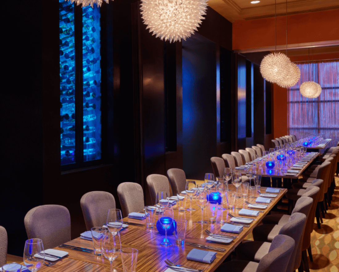 Elegant dining room with two long wooden tables set for a meal, blue glowing candle holders, modern gray chairs, and spiky white pendant lights hanging from the ceiling. Blue-lit wine racks decorate the wall.