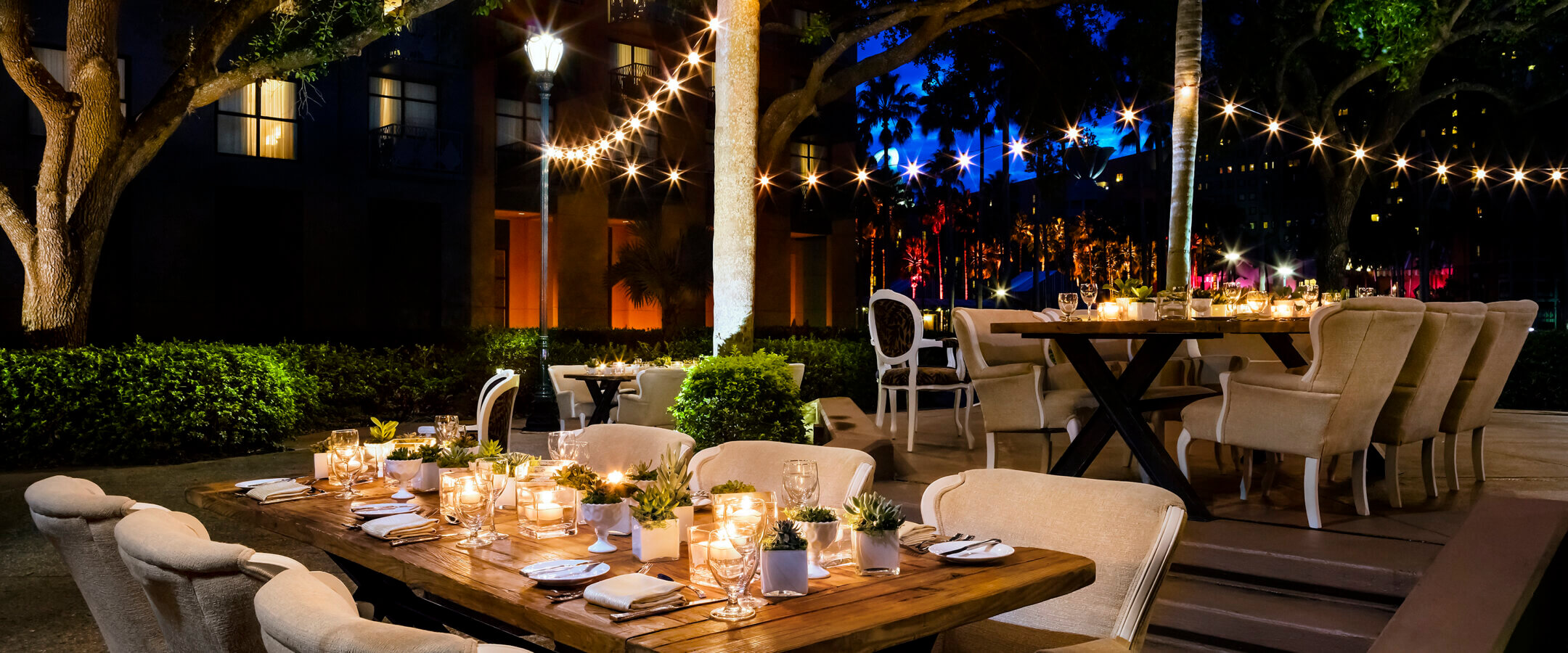 Elegant outdoor dining area at night, featuring long wooden tables with candles and small plants, beige upholstered chairs, string lights overhead, and trees and buildings in the background.